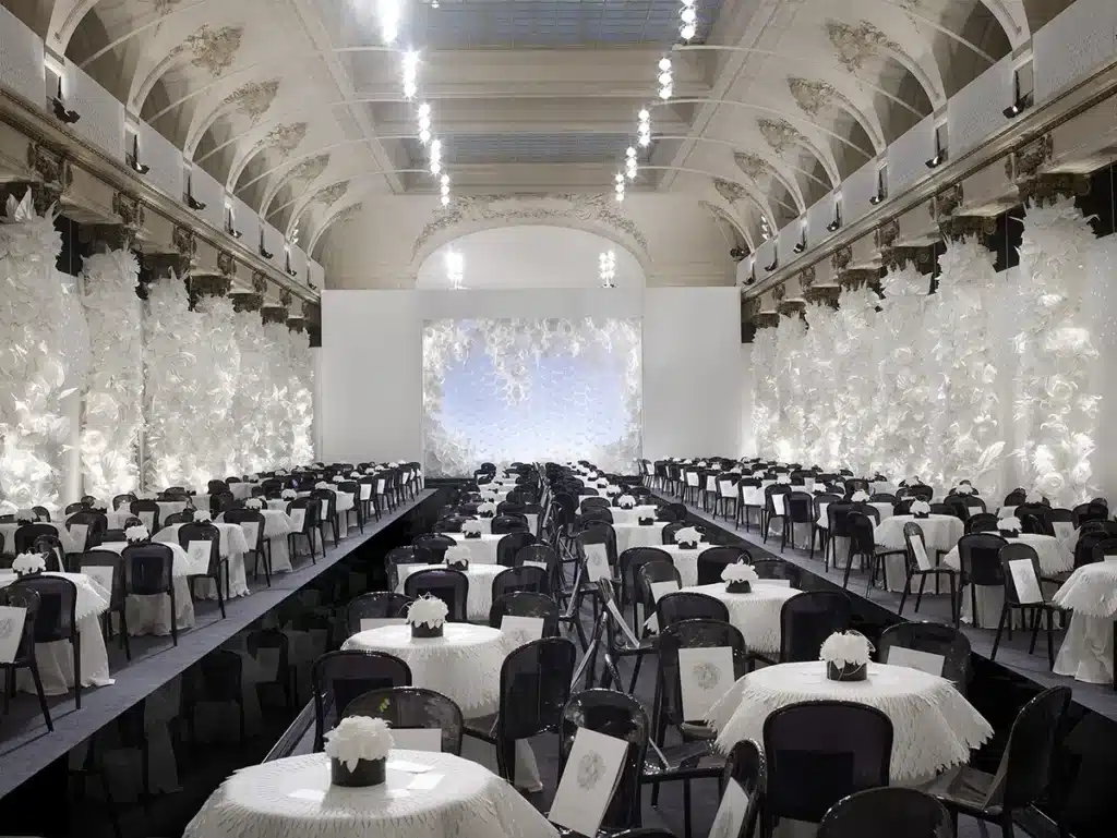 Une grande salle élégante décorée entièrement de fleurs blanches sculptées, avec de nombreuses tables rondes drapées de blanc et alignées autour d’un podium noir central, créant une ambiance raffinée et immaculée.