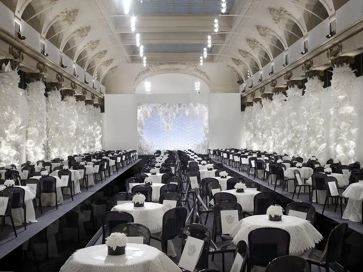Une grande salle élégante décorée entièrement de fleurs blanches sculptées, avec de nombreuses tables rondes drapées de blanc et alignées autour d’un podium noir central, créant une ambiance raffinée et immaculée.