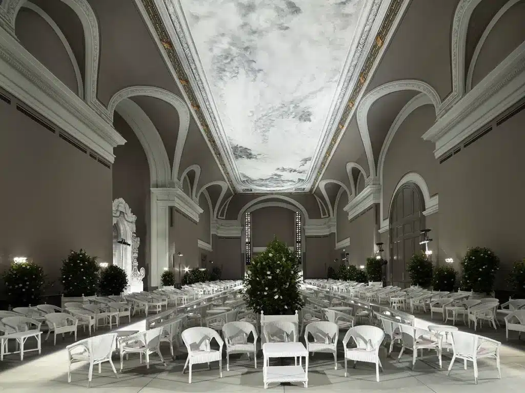 Un vaste décor intérieur aux allures de salon aristocratique, orné de grandes arches classiques et d’un plafond monumental peint façon ciel nuageux. L’espace est structuré en rangées de fauteuils et tables en rotin blanc, bordé de massifs d’orangers soigneusement disposés. L’atmosphère, élégante et lumineuse, évoque un jardin d’hiver sophistiqué, prêt à accueillir un défilé ou un événement prestigieux.