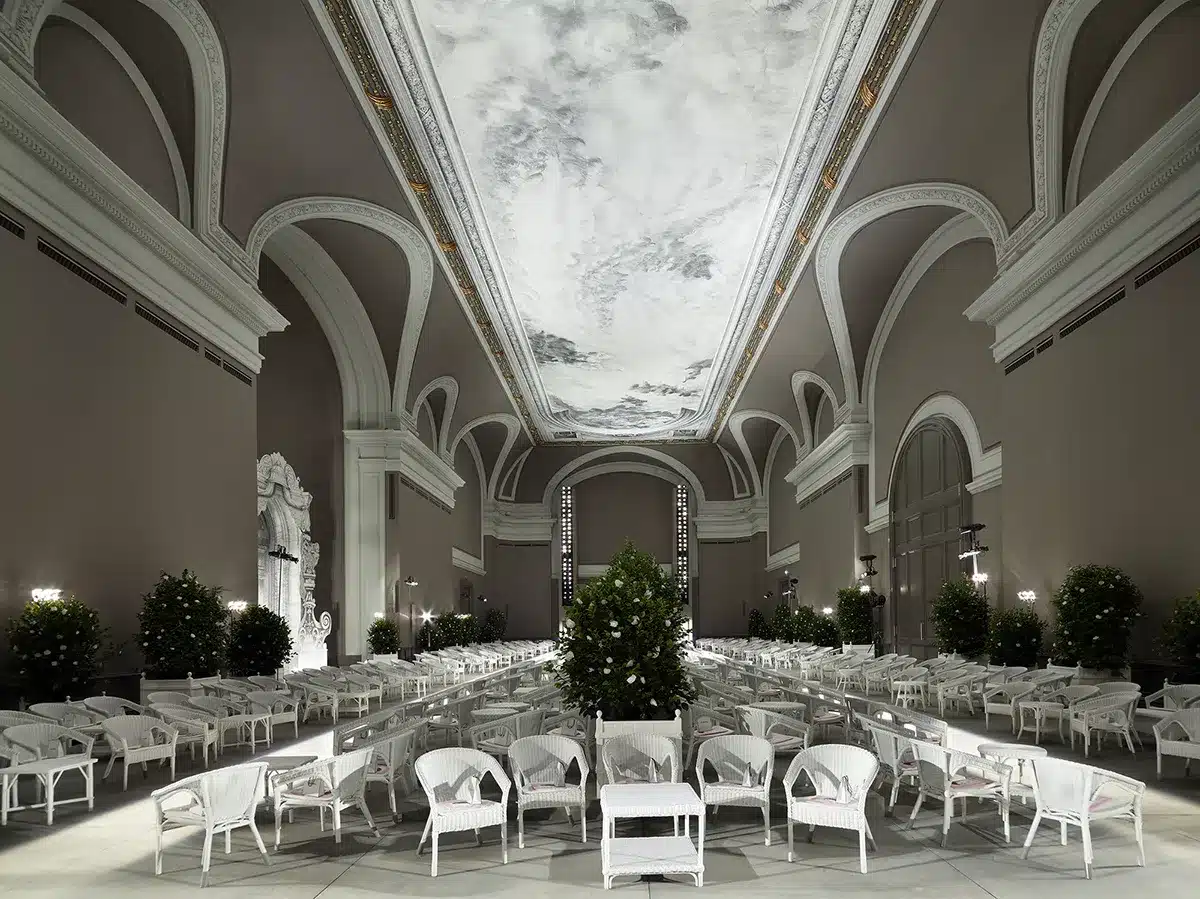 Un vaste décor intérieur aux allures de salon aristocratique, orné de grandes arches classiques et d’un plafond monumental peint façon ciel nuageux. L’espace est structuré en rangées de fauteuils et tables en rotin blanc, bordé de massifs d’orangers soigneusement disposés. L’atmosphère, élégante et lumineuse, évoque un jardin d’hiver sophistiqué, prêt à accueillir un défilé ou un événement prestigieux.