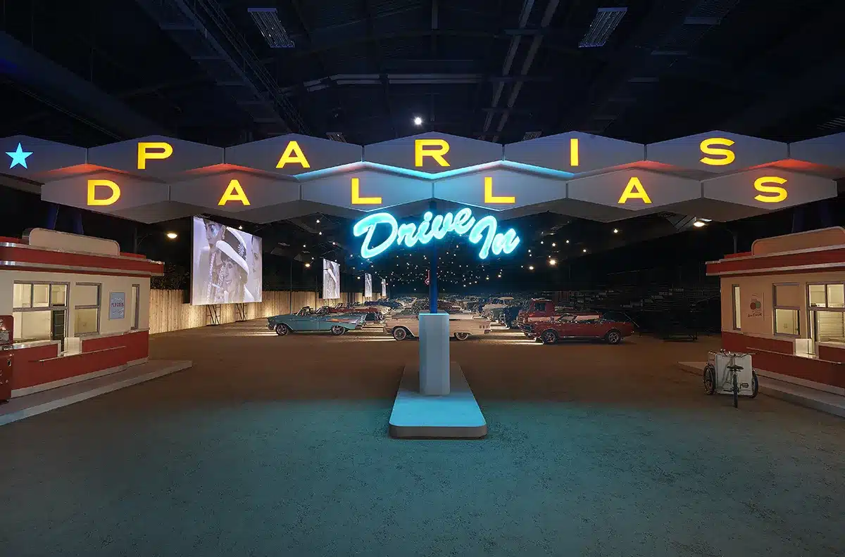 Cette image montre une reconstitution immersive d’un drive-in américain rétro, dominé par une grande enseigne lumineuse « PARIS DALLAS – Drive In ». Sous les néons bleus et jaunes, des voitures vintage sont alignées face à de grands écrans diffusant des images en noir et blanc. L’ambiance nocturne et cinématographique s’inscrit dans l’univers spectaculaire du Défilé Métiers d’Art Chanel 2013/2014 imaginé par Karl Lagerfeld, hommage à l’esthétique texane.