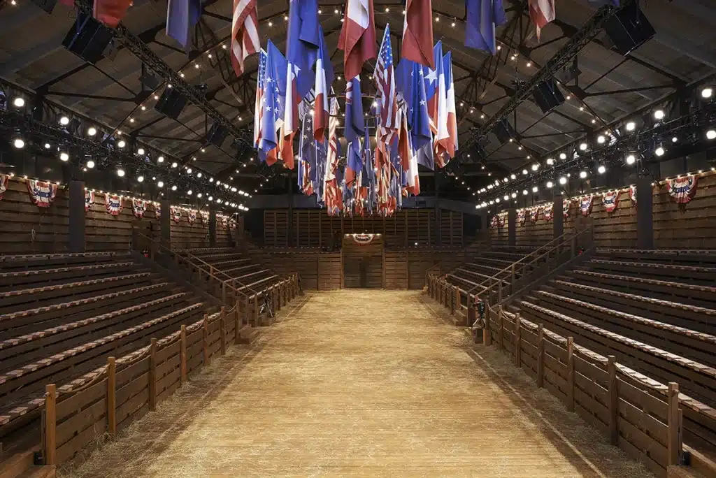 Scénographie façon rodéo pour un défilé, avec gradins en bois, sol de poussière et drapeaux américains suspendus, créant une ambiance western immersive.