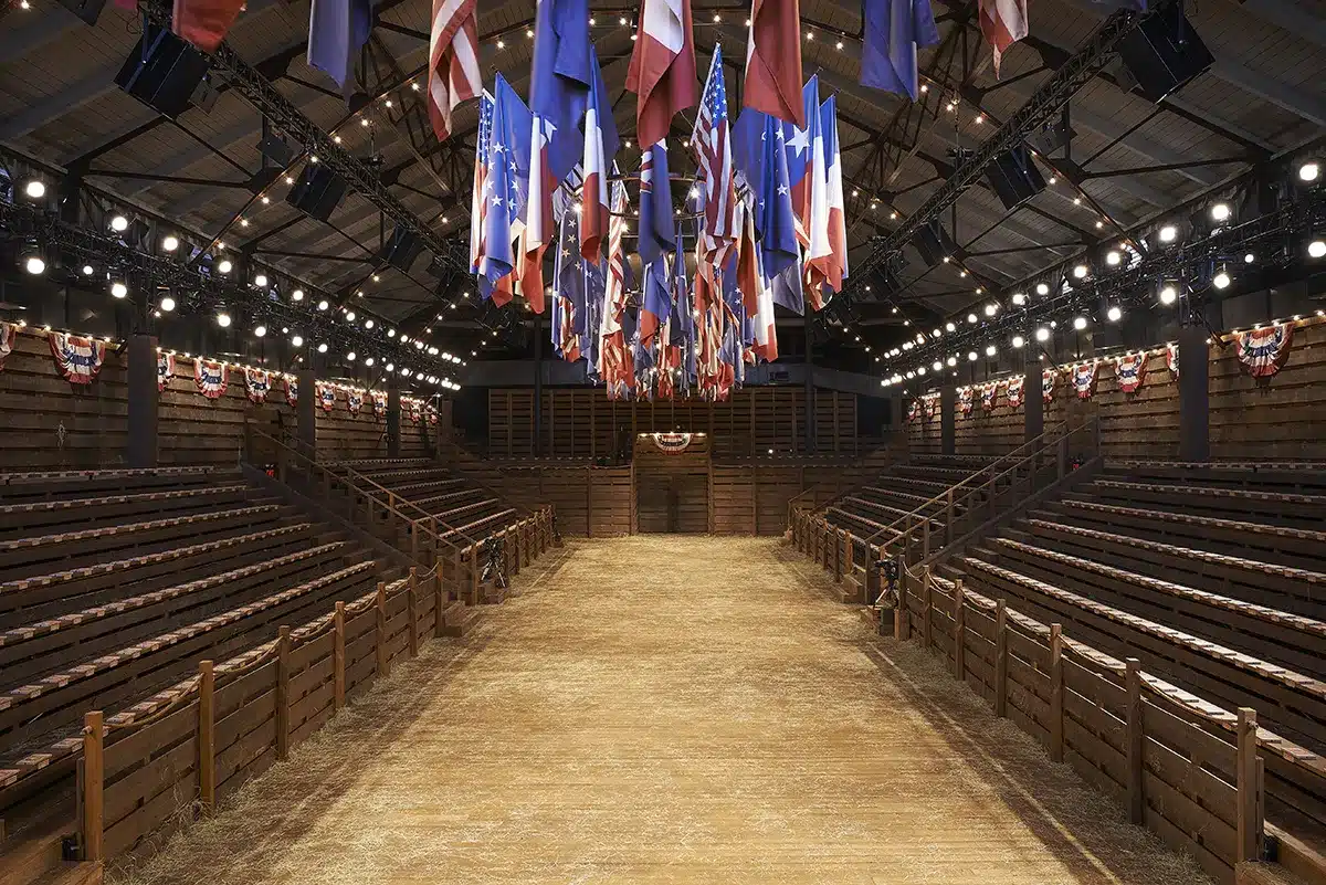 Scénographie façon rodéo pour un défilé, avec gradins en bois, sol de poussière et drapeaux américains suspendus, créant une ambiance western immersive.