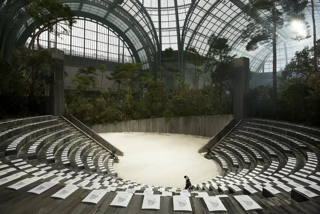 Les photos montrent une spectaculaire scénographie installée sous la verrière du Grand Palais, transformée en amphithéâtre circulaire entouré d’une forêt dense. De larges gradins en bois disposés en arc de cercle entourent une vaste arène de sable clair, tandis que des arbres et arbustes recréent un paysage naturel immersif. La lumière filtrant à travers la verrière ajoute une atmosphère poétique et majestueuse à cet espace conçu pour un défilé ou un événement artistique.
