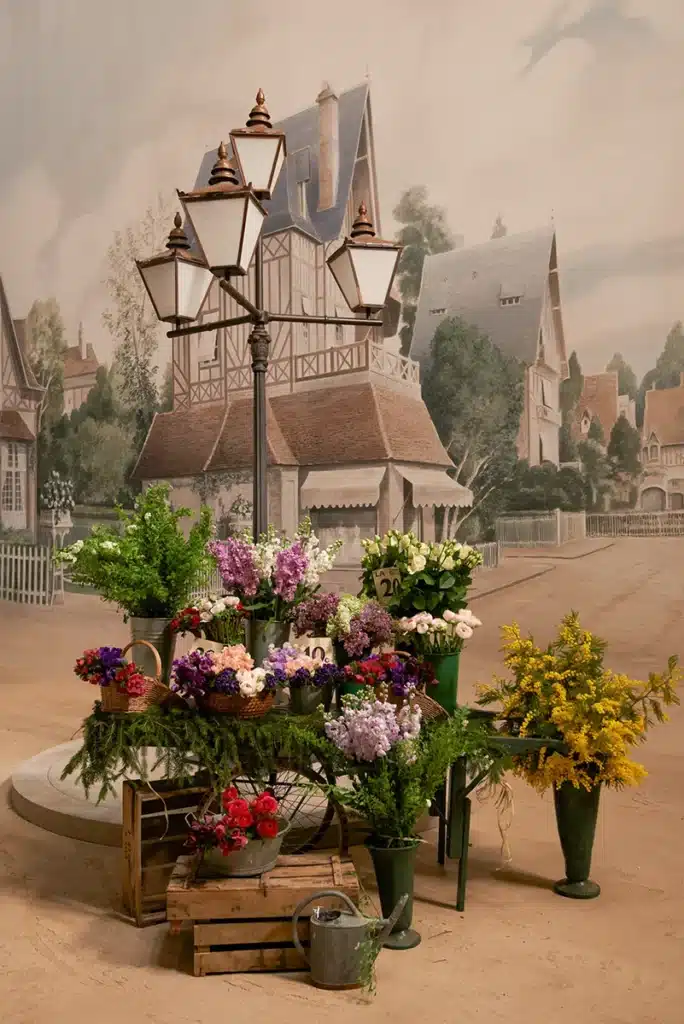 Ce décor recrée une petite ville normande du début du XXe siècle, avec ses rues calmes bordées d’arbres, ses façades à colombages, ses devantures de boutiques, son café en rotin installé en terrasse et son fleuriste aux couleurs vives. À l’intérieur, on découvre une boutique de chapeaux Gabrielle Chanel, soigneusement reconstituée avec meubles en bois, miroirs, étagères anciennes et chapeaux exposés. L’ensemble évoque une atmosphère nostalgique, élégante et artisanale, parfaite pour un film retraçant les débuts de Coco Chanel à Deauville.