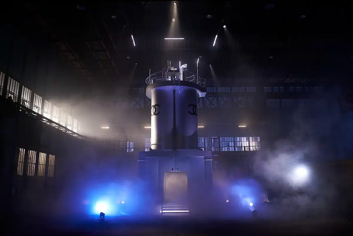 Un immense dispositif industriel futuriste, semblable à une machinerie expérimentale ou un réacteur géant, émerge au centre d’un vaste hangar plongé dans la pénombre. La structure, dominée par deux immenses cuves métalliques siglées Chanel, est accessible par une plateforme circulaire constellée de cadrans, tuyauteries et instruments techniques. Au sommet, un opérateur miniature semble surveiller la machine. L’ensemble, enveloppé de fumée et de lumières bleutées, évoque une scène à la fois scientifique, théâtrale et mystérieuse — comme un laboratoire monumental transformé en installation cinématographique.