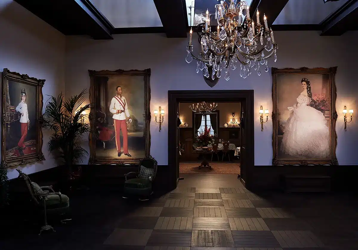 Ce décor évoque l’intérieur luxueux d’un palais impérial austro-hongrois revisité, avec de grands portraits néo-classiques encadrés d’or représentant des personnages anachroniques (enfants, Pharrell Williams, Kristen Stewart), intégrés dans des toiles peintes à la manière du XIXᵉ siècle.