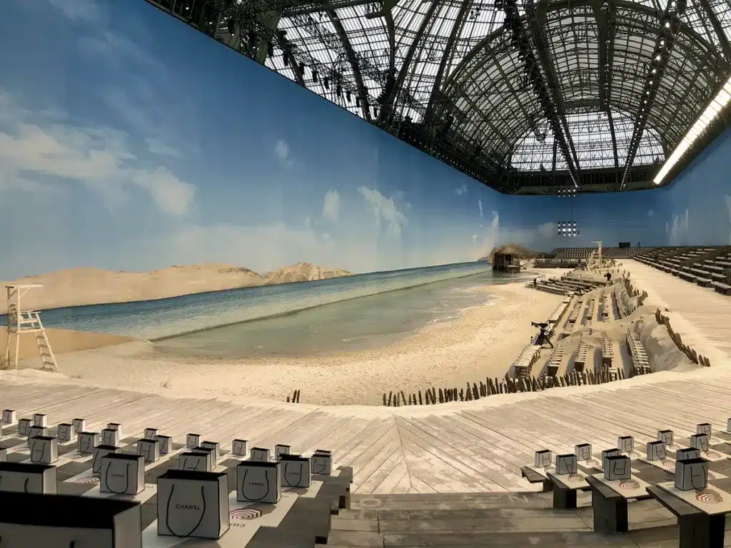 Un décor spectaculaire transformant le Grand Palais en plage immaculée, avec sable blanc, mer turquoise et ciel d’été, recréant une atmosphère de bord de mer pour un défilé Chanel immersif.