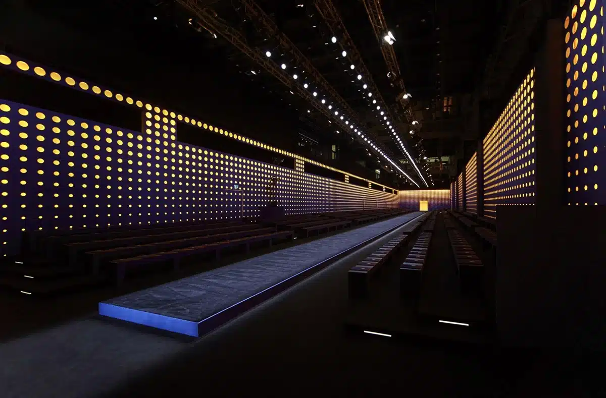 Un long podium entouré de murs sombres percés de centaines de points lumineux jaune-orangé, créant une ambiance futuriste, graphique et immersive.