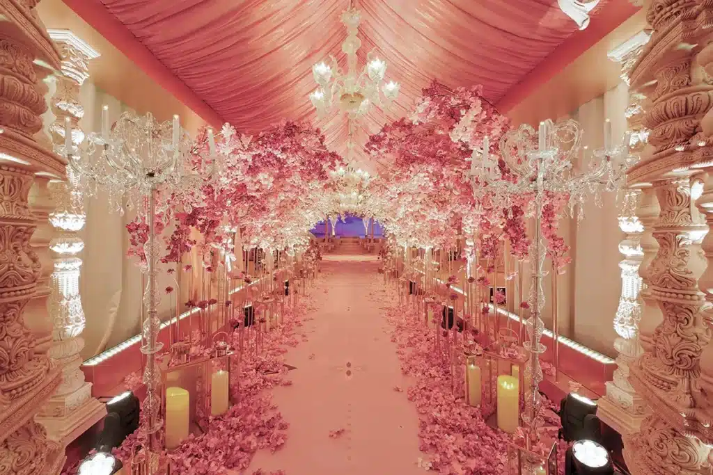 La photo montre une allée somptueusement décorée, entièrement plongée dans des tons rosés et dorés. Un plafond drapé de tissu rose descend en vagues élégantes, tandis que des lustres en cristal scintillent au-dessus de l’espace. Des arches denses de fleurs roses, rappelant des cerisiers en fleurs, enveloppent le passage et créent un tunnel floral féérique. Le sol est recouvert de pétales, et des bougeoirs, lanternes et chandeliers en cristal bordent l’allée, renforçant l’ambiance romantique et luxueuse. Au bout du chemin, quelques marches mènent à une scène ou une estrade, évoquant la préparation d’un mariage ou d’un événement très raffiné.