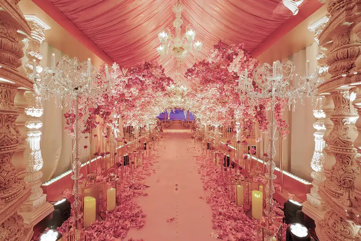 La photo montre une allée somptueusement décorée, entièrement plongée dans des tons rosés et dorés. Un plafond drapé de tissu rose descend en vagues élégantes, tandis que des lustres en cristal scintillent au-dessus de l’espace. Des arches denses de fleurs roses, rappelant des cerisiers en fleurs, enveloppent le passage et créent un tunnel floral féérique. Le sol est recouvert de pétales, et des bougeoirs, lanternes et chandeliers en cristal bordent l’allée, renforçant l’ambiance romantique et luxueuse. Au bout du chemin, quelques marches mènent à une scène ou une estrade, évoquant la préparation d’un mariage ou d’un événement très raffiné.