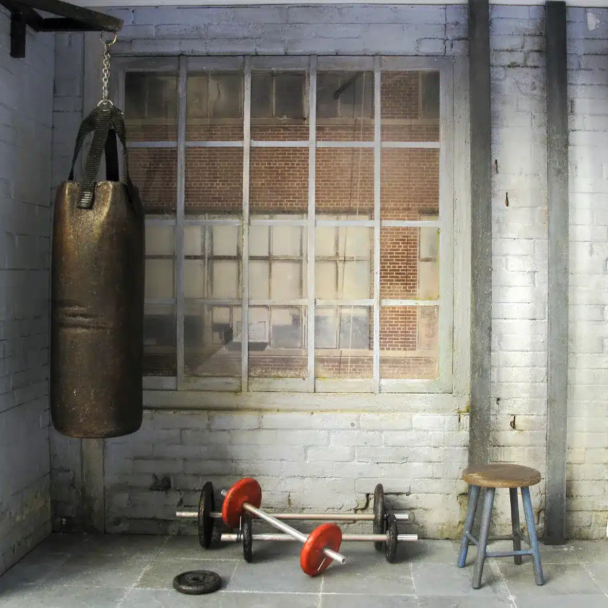 un décor miniature très réaliste avec murs en briques peintes en gris, grandes fenêtres industrielles, punching-ball usé, poids et haltères rouges, banc simple et tabouret en bois. L’ambiance est brute, urbaine et authentique, comme un petit gym de quartier.