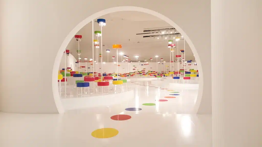 Cette image montre un décor immersif : une vaste salle blanche dans laquelle une ouverture circulaire révèle un espace rempli de colonnes lumineuses surmontées de disques colorés (rouge, jaune, bleu, vert). L’ensemble crée une ambiance ludique et presque futuriste, comme une forêt graphique composée de points lumineux et de volumes géométriques. Le sol immaculé renforce l’effet d’expansion visuelle et met en valeur la répétition rythmique des couleurs et des formes.