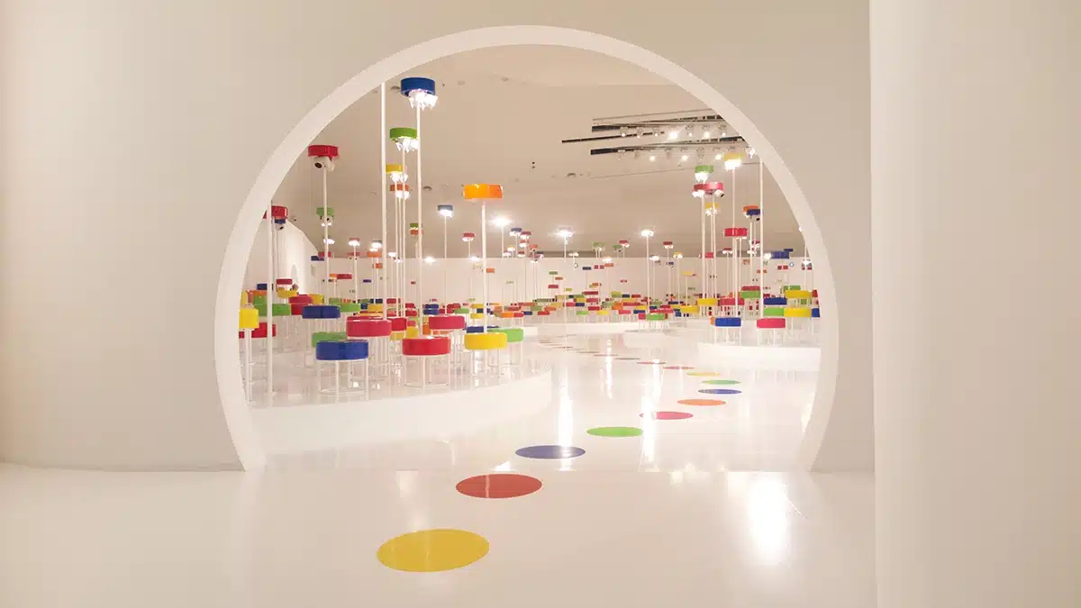 Cette image montre un décor immersif : une vaste salle blanche dans laquelle une ouverture circulaire révèle un espace rempli de colonnes lumineuses surmontées de disques colorés (rouge, jaune, bleu, vert). L’ensemble crée une ambiance ludique et presque futuriste, comme une forêt graphique composée de points lumineux et de volumes géométriques. Le sol immaculé renforce l’effet d’expansion visuelle et met en valeur la répétition rythmique des couleurs et des formes.