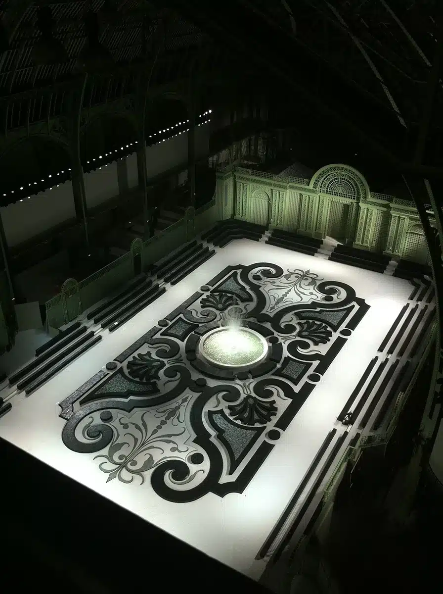 Une vaste mise en scène au Grand Palais, avec un sol blanc orné d’un immense motif baroque noir et gris, centré autour d’une fontaine lumineuse, encadré de gradins et d’une façade décorative vert pâle.