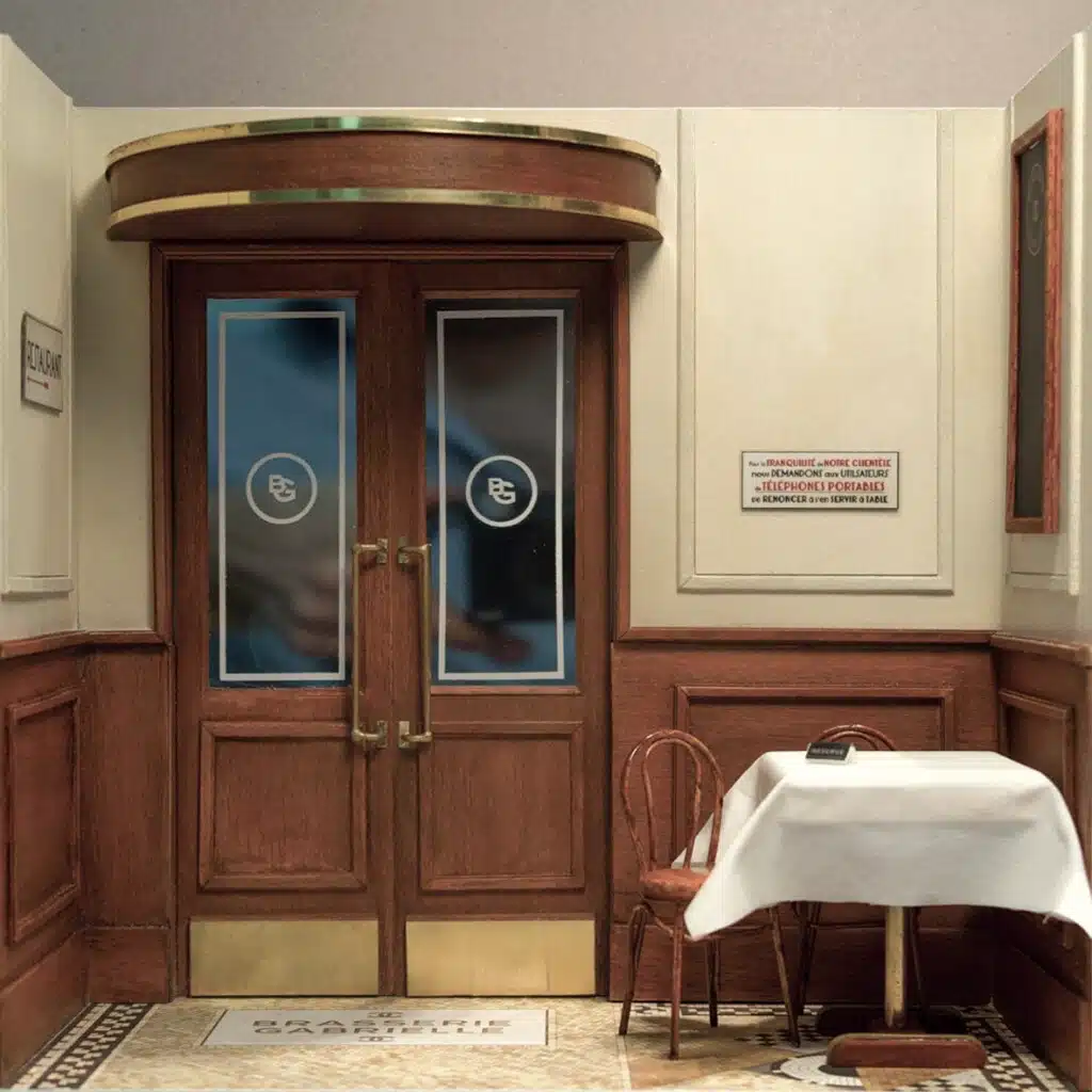 Maquette d'Une vitrine Chanel transformée en entrée de brasserie parisienne, avec de grandes portes en bois sombre et laiton, des mannequins élégants comme des clientes chic, et des chaises empilées ou dressées autour d’une petite table, créant une ambiance Paris rétro, raffinée et chaleureuse.