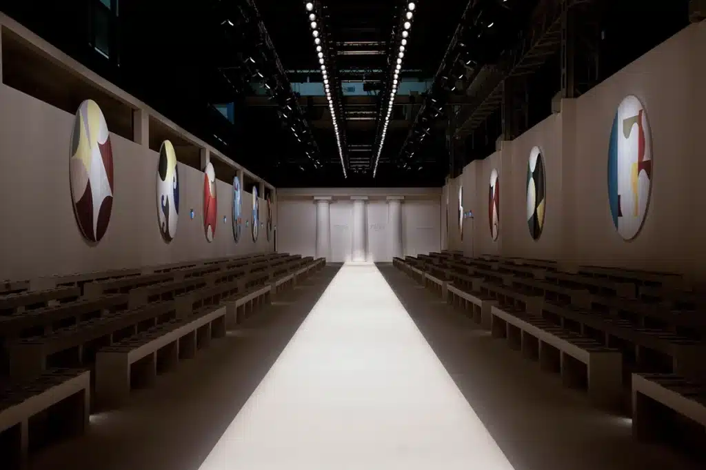 Un podium épuré s’étire au centre d’un décor blanc minimaliste, encadré par de longues gradins rectilignes. Les murs latéraux sont rythmés par de grands médaillons colorés, aux motifs abstraits et graphiques qui apportent une touche artistique à l’espace. Au fond, trois colonnes sobres structurent l’entrée du défilé, où apparaît discrètement le nom FENDI. L’ensemble offre une atmosphère à la fois moderne, calme et architecturale, mettant en valeur les silhouettes du show.
