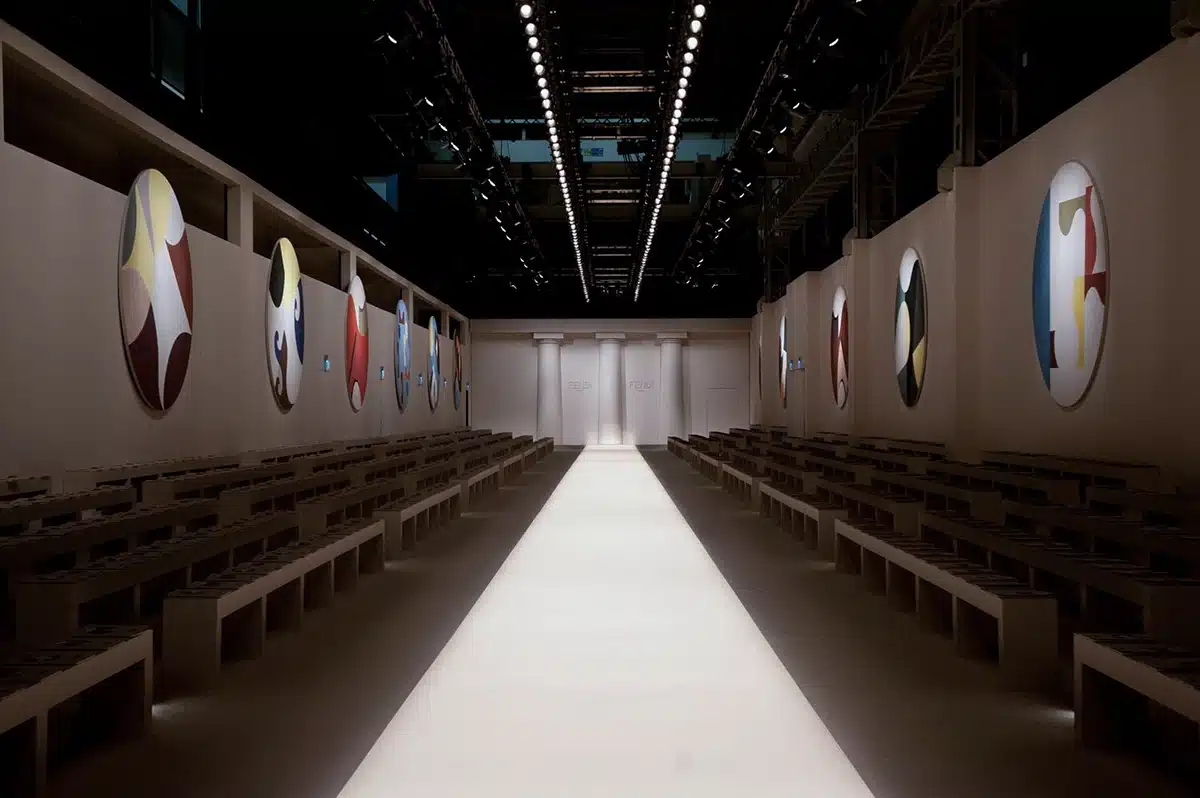 Un podium épuré s’étire au centre d’un décor blanc minimaliste, encadré par de longues gradins rectilignes. Les murs latéraux sont rythmés par de grands médaillons colorés, aux motifs abstraits et graphiques qui apportent une touche artistique à l’espace. Au fond, trois colonnes sobres structurent l’entrée du défilé, où apparaît discrètement le nom FENDI. L’ensemble offre une atmosphère à la fois moderne, calme et architecturale, mettant en valeur les silhouettes du show.