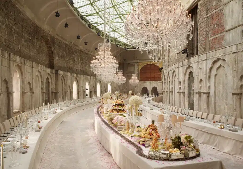 Cette photo montre une immense salle transformée en banquet somptueux. Dans un décor de murs patinés d’inspiration indo-moghole, une longue table courbe est richement dressée : compositions florales, pâtisseries et pièces montées se succèdent sous une multitude de lustres en cristal suspendus. L’ensemble crée une atmosphère opulente, théâtrale et raffinée, contrastant avec l’aspect volontairement vieilli des architecture.