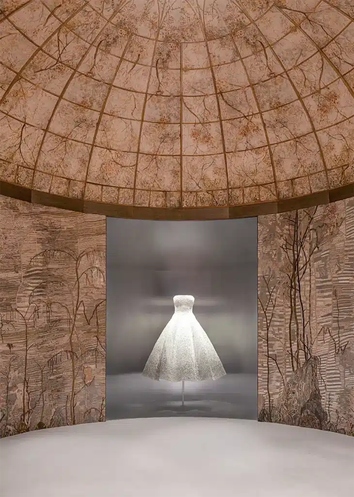 Espace d’exposition immersif mettant en valeur deux robes haute couture finement brodées, chacune présentée sous vitrine dans une alcôve courbe décorée de motifs floraux. Les murs et le plafond forment une coupole tapissée d’illustrations végétales délicates d'Eva Jospin, créant une atmosphère chaleureuse et artisanale qui souligne le raffinement des pièces exposées.