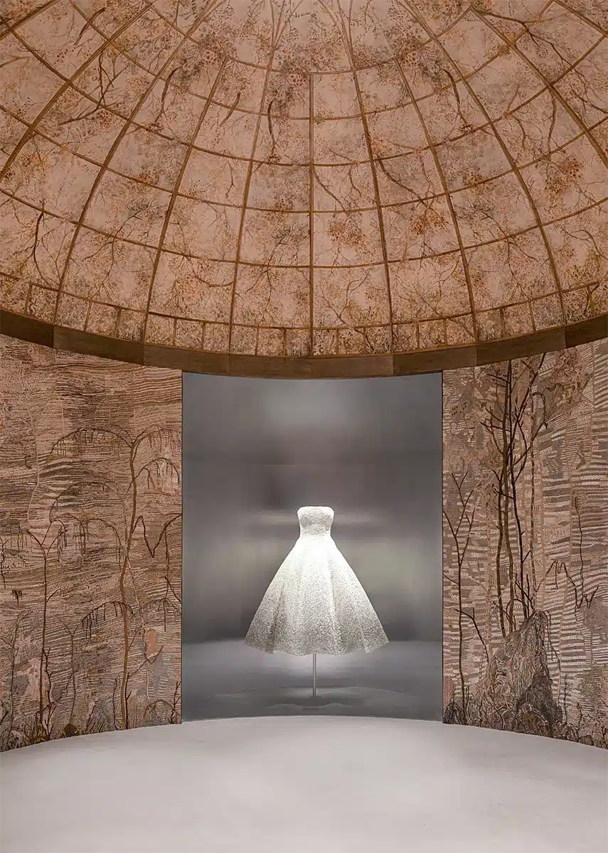 Espace d’exposition immersif mettant en valeur deux robes haute couture finement brodées, chacune présentée sous vitrine dans une alcôve courbe décorée de motifs floraux. Les murs et le plafond forment une coupole tapissée d’illustrations végétales délicates d'Eva Jospin, créant une atmosphère chaleureuse et artisanale qui souligne le raffinement des pièces exposées.