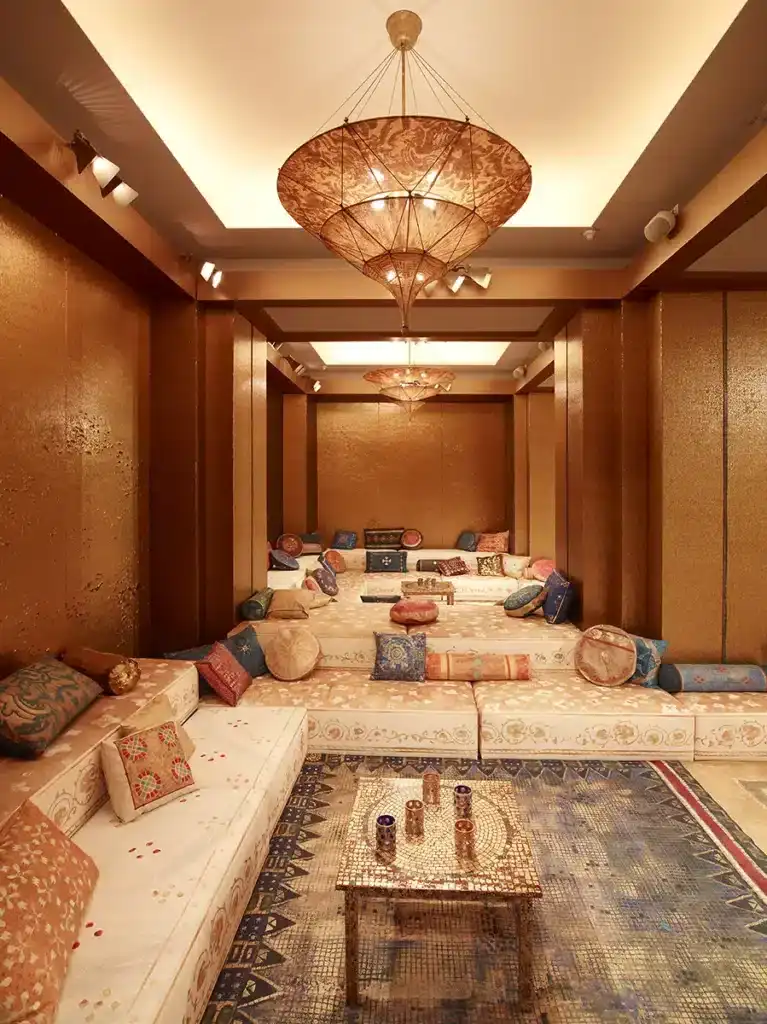 Un salon luxueux impérial byzantin, entièrement drapé de panneaux dorés aux textures scintillantes. L’espace est structuré en gradins tapissés de larges banquettes crème, ornées de motifs brodés et abondamment recouvertes de coussins richement décorés : velours, brocarts, couleurs safran, bleu nuit, or et rouge. De grands tapis mosaïqués couvrent le sol et rappellent l’esthétique persane. Au plafond, des lustres en forme de lanternes tissées, suspendus comme des voiles, diffusent une lumière chaude et tamisée. L’ensemble crée une atmosphère opulente, intime et chaleureuse, évoquant un boudoir oriental contemporain, élégant et profondément immersif.