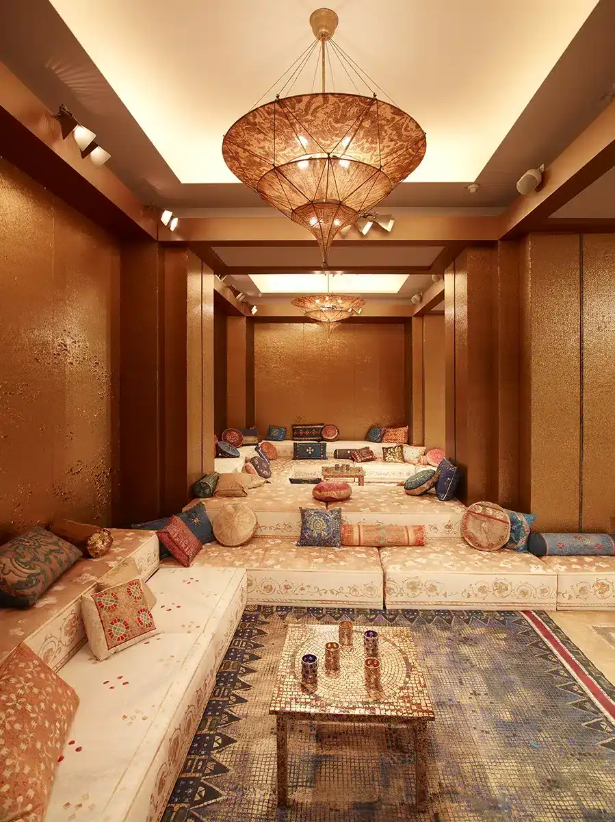 Un salon luxueux impérial byzantin, entièrement drapé de panneaux dorés aux textures scintillantes. L’espace est structuré en gradins tapissés de larges banquettes crème, ornées de motifs brodés et abondamment recouvertes de coussins richement décorés : velours, brocarts, couleurs safran, bleu nuit, or et rouge. De grands tapis mosaïqués couvrent le sol et rappellent l’esthétique persane. Au plafond, des lustres en forme de lanternes tissées, suspendus comme des voiles, diffusent une lumière chaude et tamisée. L’ensemble crée une atmosphère opulente, intime et chaleureuse, évoquant un boudoir oriental contemporain, élégant et profondément immersif.