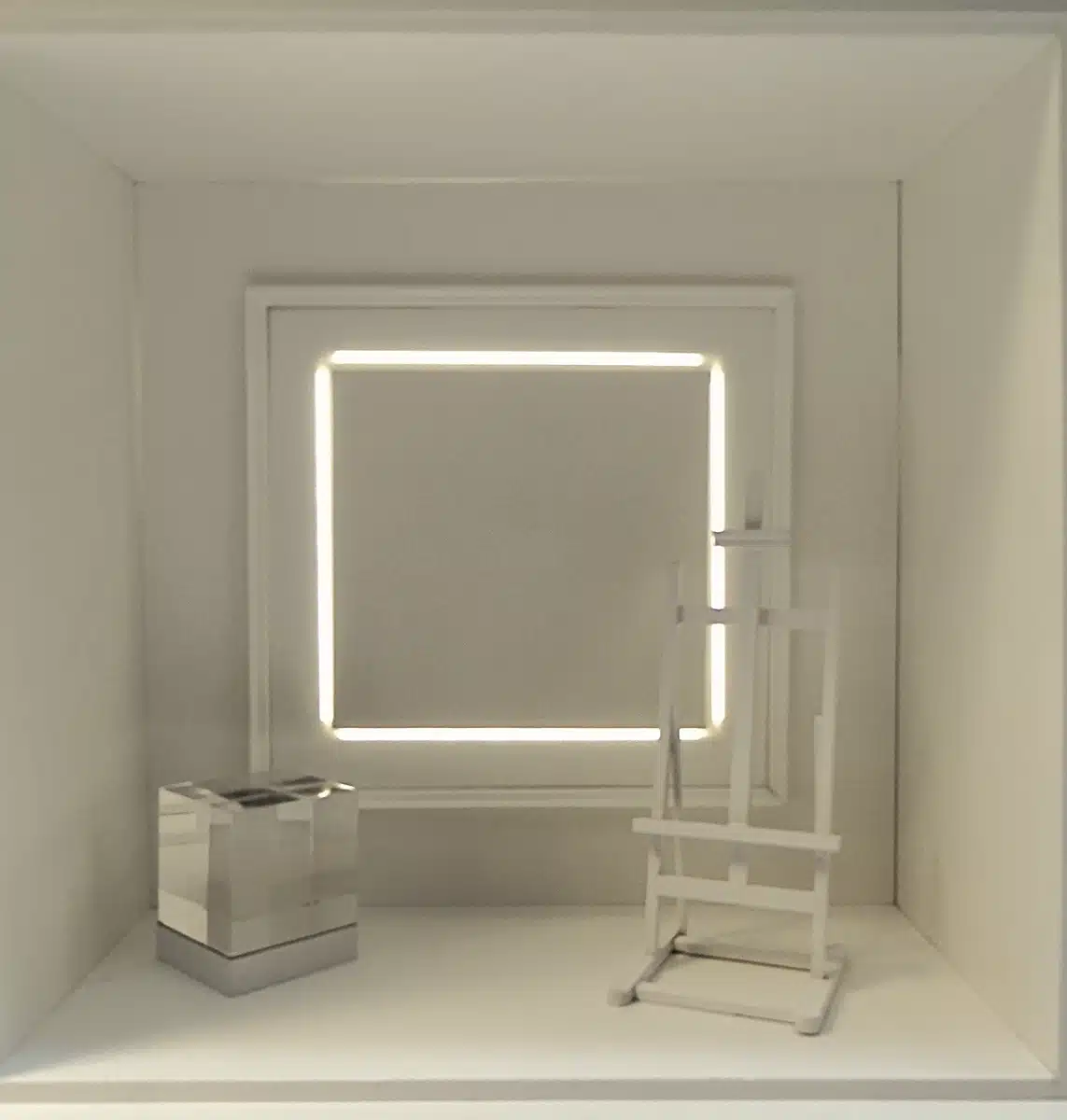 Ces vitrines jouent sur une esthétique minimaliste et lumineuse : un cadre lumineux géométrique structure l’espace, tandis que des volumes transparents ou miroir — cubes, colonnes, socles — mettent en valeur les produits. L’ensemble, très épuré et monochrome, crée une atmosphère contemporaine et presque muséale, où la lumière devient l’élément central de scénographie. Les mannequins, habillés de silhouettes colorées et graphiques, contrastent avec ce décor sobre, renforçant l’effet de mise en scène artistique.