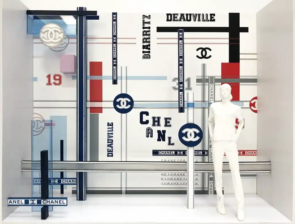 maquettes présentent un univers graphique très structuré, composé de lignes parallèles, bandes colorées et typographies inspirées de plans urbains et de signalétique sportive. Les couleurs principales — rouge, bleu, noir, or et gris — encadrent un mannequin blanc minimaliste placé au centre, mettant en scène l’espace comme une composition graphique en trois dimensions. Les éléments emblématiques de la maison Chanel (logo CC, chiffre 5, numéros 19, 31) surgissent sous forme de blocs en relief. L’ensemble évoque un décor moderne, rythmé et architectural, à la fois ludique et élégant, déployé ensuite en vitrine réelle où les mannequins portent les silhouettes Chanel devant cette cartographie stylisée.