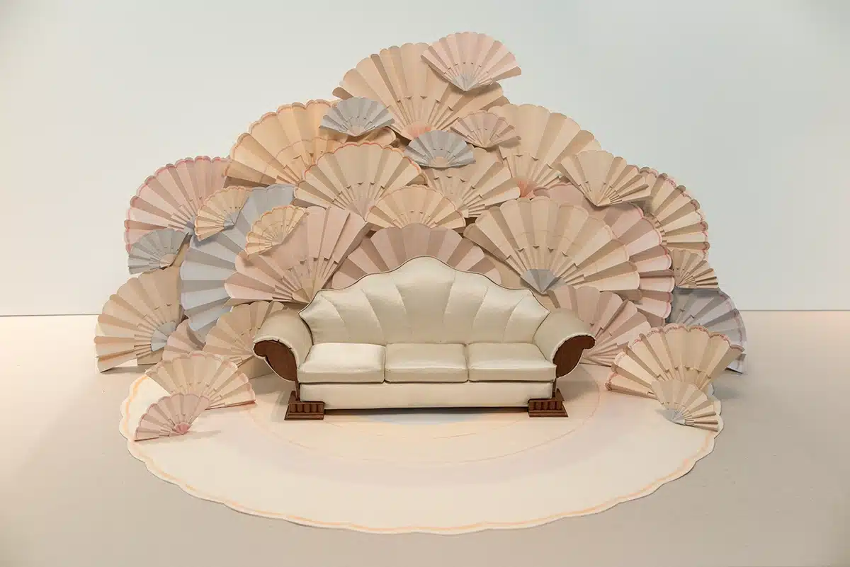 Cette scénographie se déploie comme un décor délicat et théâtral : un canapé aux lignes vintage est enveloppé d’un mur de grands éventails en papier, déclinés en tons pastel poudrés — beige, rose, pêche et touches de gris. Superposés et évasés, ils créent un halo presque floral autour de la scène