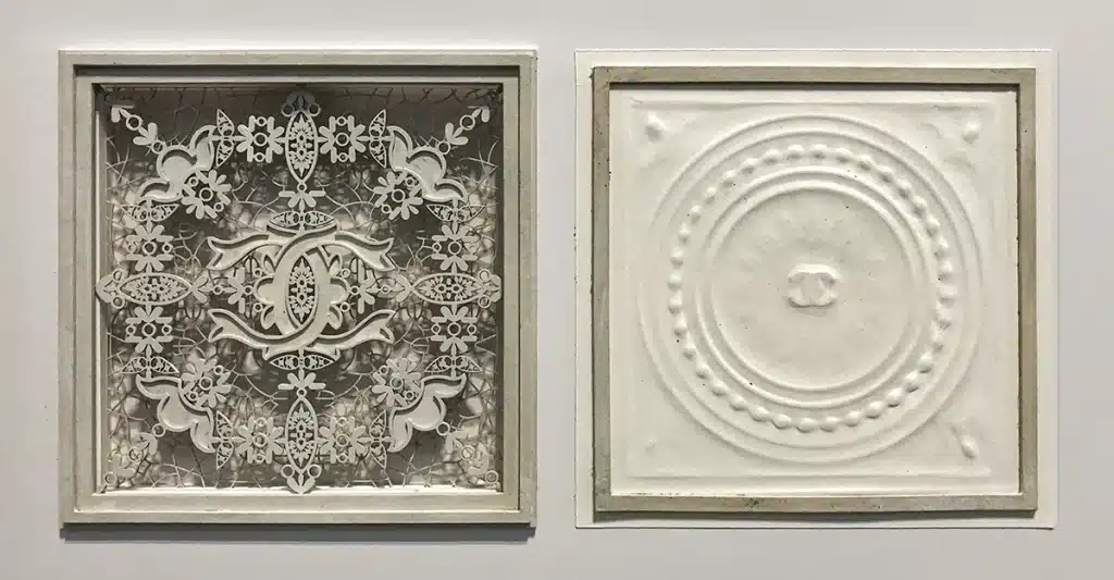 Une composition entièrement blanche et délicate inspirée d’une dentelle sculptée : un grand panneau encadré présente un motif ornemental ajouré très fin, organisé autour du double C iconique de Chanel. Les éléments découpés, superposés et légèrement en relief créent un effet de broderie luxueuse, mis en valeur par un fond texturé et un éclairage doux. L’ensemble évoque l’élégance classique et la précision artisanale propre à la maison Chanel.