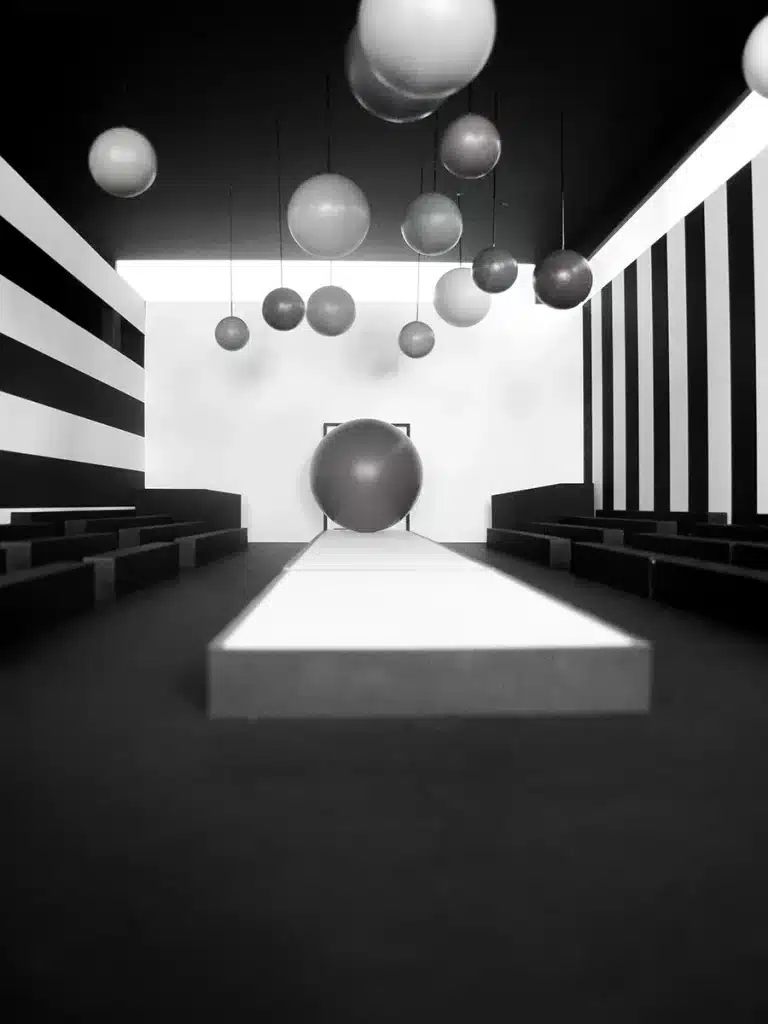 Une scénographie monochrome et très graphique, organisée autour d’un podium blanc encadré de gradins noirs. D’immenses sphères suspendues – mates, grises et argentées – flottent dans l’espace comme un nuage géométrique, tandis qu’une sphère géante marque le fond du défilé. Les murs latéraux, habillés de larges rayures noires et blanches, renforcent l’effet optique et la profondeur. L’ensemble crée une ambiance futuriste, minimaliste et immersive, jouant sur les volumes flottants et les contrastes.