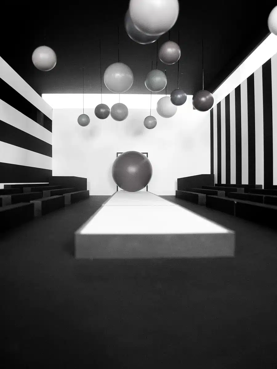 Une scénographie monochrome et très graphique, organisée autour d’un podium blanc encadré de gradins noirs. D’immenses sphères suspendues – mates, grises et argentées – flottent dans l’espace comme un nuage géométrique, tandis qu’une sphère géante marque le fond du défilé. Les murs latéraux, habillés de larges rayures noires et blanches, renforcent l’effet optique et la profondeur. L’ensemble crée une ambiance futuriste, minimaliste et immersive, jouant sur les volumes flottants et les contrastes.