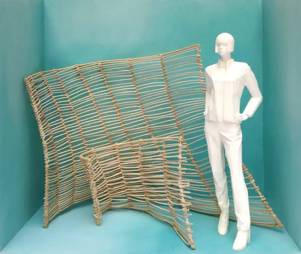 Une scénographie minimaliste présente un mannequin blanc stylisé, aux formes angulaires, posé dans un cube turquoise. À côté de lui, une grande structure organique en vannerie légère, composée de tiges fines tressées, crée une forme ondulante et sculpturale.
