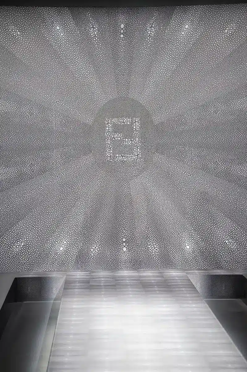 L'image montre un podium minimaliste et futuriste, entièrement décliné dans une palette de gris métallisés. Le fond de scène est occupé par un immense panneau imprimé d’un motif galuchat, formant un effet visuel radiant qui converge vers un centre où apparaît un logo Fendi stylisé. Le podium reprend ce même traitement texturé, créant une continuité visuelle avec le mur. Les gradins, géométriques et épurés, encadrent le défilé dans une ambiance très architecturale et monolithique. L’ensemble évoque un univers luxe, presque clinique, où la matière galuchat devient un motif graphique hypnotique.