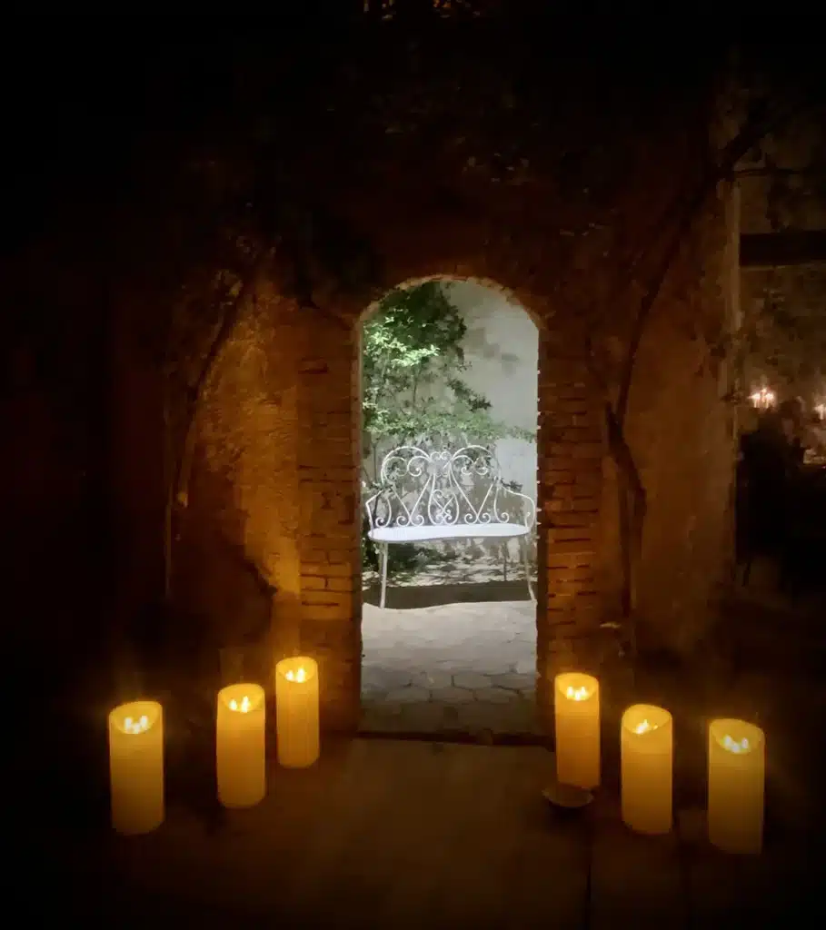 Une ambiance élégante et romantique dans une grange en pierre : longues tables nappées de blanc, fleurs pastel abondantes, chandeliers et lustres en cristal. La décoration mêle charme ancien et poésie bucolique, avec bougies, verdure, vaisselle raffinée et coins intimistes éclairés à la lueur chaude, créant une atmosphère chaleureuse et festive.